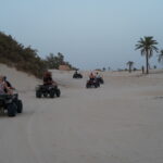 Le Sahara Sud Tunisien est un monde à part. Nous avons tous déjà entendu parler de ses dunes dorées, de ses oasis secrètes et de sa lumière singulière. Mais le découvrir en vrai, c’est autre chose. Imaginez-vous, le soir, assis confortablement près d’un feu de camp, écoutant le bruit discret du vent dans le désert, tandis que le soleil, rougi par la journée, cède doucement la place à un ciel scintillant d’étoiles. Ce n’est pas juste un voyage, c’est une aventure humaine, une immersion totale dans un univers qui semble hors du temps. Au-delà des clichés, le tourisme en Tunisie se révèle beaucoup plus profond et fascinant que de simples vacances au bord de la mer. Le Sahara nous invite à une quête d’authenticité, à la rencontre de traditions ancestrales, à la découverte de paysages indescriptibles et de cultures rurales préservées. Explorer la Culture du Sahara Sud Tunisien : Rencontres et Traditions Ancestrales Le Sahara Sud Tunisien offre un mélange surprenant de traditions, de croyances et de modes de vie qui ont su traverser les siècles sans perdre leur âme. Nous considérons cette région comme un véritable carrefour culturel, où l’on découvre des villages berbères accrochés au désert, des artisans passionnés, et des familles qui perpétuent encore aujourd’hui des coutumes millénaires. C’est en pénétrant dans ces espaces que l’on mesure la richesse culturelle de la Tunisie profonde, celle qui ne s’expose pas toujours sur les cartes postales. En vous aventurant dans les villages fortifiés – appelés ksour – vous aurez l’impression de remonter le temps. Ces constructions en terre ocre, protégées autrefois par des remparts solides, sont aujourd’hui des témoins vivants de l’histoire locale. Parcourir leurs ruelles étroites, admirer la façon dont les habitants ont su dompter un environnement exigeant, c’est comprendre la résilience d’un peuple qui ne cesse de s’adapter. Vous pourrez échanger avec les habitants, goûter aux saveurs de la cuisine traditionnelle, contempler les tapis faits à la main et parfois vous arrêter devant un tisserand ou un potier, perpétuant avec fierté un savoir-faire transmis de génération en génération. L’accueil chaleureux que vous réserve cette région est aussi l’une des grandes richesses du Sahara Sud Tunisien. Ici, on prend le temps. On vous invite à vous asseoir, à partager un thé menthe brûlant, à écouter une histoire racontée par un ancien. Ce sont ces moments simples, mais précieux, qui rendent un séjour dans le désert inoubliable. Merveilles Paysagères : Dunes, Oasis et Lumières du Désert Le Sahara Sud Tunisien n’est pas qu’une étendue sans fin de sable. Bien sûr, on est d’abord impressionnés par ces dunes monumentales, changeant de couleur au fil de la journée, passant du jaune pâle à l’orange vif, jusqu’au rose pastel lorsque le soleil se couche. Marcher dans le sable doux, sentir le vent léger qui modèle sans cesse le relief, c’est une expérience apaisante, presque méditative. Nous recommandons de prendre le temps d’observer les jeux de lumière, les ombres portées, ces détails infimes qui forment un tableau vivant. Mais le désert réserve aussi des surprises inattendues. Imaginez arriver, après une longue route, à une oasis. Soudain, la sécheresse laisse place au vert des palmiers, au murmure de l’eau qui s’écoule doucement dans les rigoles d’irrigation, aux jardins potagers qui prospèrent malgré tout. Ces points de verdure surgissent tels des miracles au milieu du néant. Vous y trouverez parfois des sources thermales, des bassins où se rafraîchir, des villages paisibles où l’on vit essentiellement de l’agriculture. Les circuits dans le désert proposés par des prestataires locaux permettent de découvrir des joyaux comme Ksar Ghilane, une oasis emblématique où l’on peut se baigner dans une source naturelle au milieu des dunes. Pour en savoir plus, n’hésitez pas à consulter ce lien. Les amateurs de photographie seront comblés. Le désert inspire, il pousse à se concentrer sur les formes et les couleurs. C’est un terrain de jeu inépuisable pour capturer la beauté brute de la nature. Du lever du soleil qui fait danser les ombres sur le sable aux silhouettes gracieuses des dromadaires se détachant sur l’horizon, chaque instant est un cliché potentiel. Activités, Circuits et Excursions : Expériences Intenses et Sensations Fortes Découvrir le Sahara Sud Tunisien ne se limite pas à regarder le paysage, c’est aussi le vivre, le ressentir et le parcourir. Nous savons qu’au-delà de la contemplation, vous souhaitez peut-être plus d’action. Vous avez de la chance, car la région regorge d’expériences insolites, mêlant aventure et authenticité. Les circuit et excursion dans le désert se déclinent de mille façons : en 4x4, à dos de dromadaire, en quad ou même à pied, en mode trek, si vous êtes amateurs de randonnées sportives. Vous pouvez choisir un parcours court, juste pour quelques heures, ou opter pour un itinéraire plus long, étalé sur plusieurs jours, avec des bivouacs sous les étoiles. Passer une nuit sous une tente, au cœur du silence du désert, est une expérience à part. L’air est pur, le ciel incroyablement clair, et vous vous endormez en écoutant le crépitement du feu de camp. Pour les plus audacieux, il est possible de tester le sandboarding, une sorte de surf sur les dunes, ou de s’aventurer dans les canyons rocheux qui cachent parfois des gorges sculptées par les pluies rares mais violentes. Les levers et couchers de soleil constituent aussi des moments forts. Observer le désert au petit matin, quand la lumière est douce et que la température n’a pas encore grimpé, procure une sensation de calme absolu. Une excursion dans le Sahara Sud Tunisien, c’est également l’occasion de découvrir des sites emblématiques de la région. Pensez à la fameuse oasis de Chebika, perdue dans les montagnes, ou encore à Douz, appelée la "porte du désert". Cette petite ville dynamique propose un marché animé où l’on peut trouver des produits locaux, des épices, des étoffes. De là, on part facilement pour des safaris-photos, des promenades à dromadaire ou des sorties en 4x4, histoire de s’immerger pleinement dans l’immensité du Sahara. Le Sahara Sud Tunisien : Un Patrimoine Historique et Cinématographique Le Sahara Sud Tunisien a non seulement inspiré les voyageurs au fil des siècles, mais il a également servi de décor à de nombreux tournages. Les cinéphiles seront ravis de découvrir les lieux où ont été filmées certaines scènes mythiques. Des productions hollywoodiennes ont posé leurs caméras dans ces paysages sauvages. Par exemple, la région de Tataouine a donné son nom à une planète dans la célèbre saga Star Wars. Les amateurs de science-fiction y trouveront un certain charme, en contemplant ces décors naturels qui semblent sortis d’un film. Mais le désert est avant tout un livre d’histoire. Les vestiges romains, les anciennes routes caravanières, les greniers fortifiés (appelés ghorfas), témoignent de l’importance stratégique et commerciale de la région autrefois. Les ksour, avec leurs multiples étages, servaient à stocker les denrées, mais aussi à protéger les biens des pillards. Les découvrir, c’est s’offrir une plongée dans le passé, à une époque où les caravaniers traversaient le Sahara pour échanger épices, or, étoffes précieuses ou dattes savoureuses. Gastronomie et Saveurs du Désert : Déguster la Tunisie Authentique Un voyage dans le Sahara Sud Tunisien, c’est aussi une aventure culinaire. Nous conseillons de vous laisser tenter par les spécialités locales, élaborées avec des produits simples mais savoureux. Goûtez aux dattes fraîches, charnues et sucrées, récoltées sur place. Dégustez un couscous parfumé, préparé avec des légumes du cru, de la viande de mouton ou de chameau, et surtout beaucoup de patience. Le pain cuit dans le sable est une curiosité à ne pas manquer. Les épices locales – cumin, coriandre, paprika – rehaussent les plats d’une touche envoûtante. Le thé à la menthe, omniprésent, constitue un rituel de bienvenue. On vous l’offrira dès votre arrivée, et c’est un véritable plaisir que de le siroter, installé confortablement, en observant le rythme lent et serein du désert. La gastronomie saharienne est le reflet de son environnement : des saveurs franches, peu d’artifices, mais une authenticité qui donne envie d’en reprendre encore. Choisir le Moment Idéal et Planifier Son Séjour dans le Sahara Sud Tunisien Même si le désert a son charme toute l’année, il est plus agréable de le découvrir aux saisons douces. Le printemps et l’automne offrent des températures raisonnables, idéales pour les balades et les excursions. En été, la chaleur peut être écrasante, mais certains voyageurs apprécient malgré tout cette atmosphère presque irréelle, où la lumière est intense et les ombres courtes. En hiver, les nuits sont fraîches, mais les journées souvent clémentes, un compromis intéressant pour ceux qui recherchent le calme et la tranquillité. Il est recommandé de choisir des guides et des agences locales, qui connaissent parfaitement le terrain et les particularités du climat. Ils vous proposeront des circuits adaptés à vos envies, votre niveau de confort, votre budget. Vous pourrez ainsi opter pour un hébergement simple, une tente dans le désert, ou un campement plus confortable, équipé de lits douillets et d’un espace de restauration convivial. N’hésitez pas à solliciter un devis personnalisé pour préparer votre séjour dans le Sahara Sud Tunisien en consultant ce lien de contact. C’est une étape utile, afin de définir précisément le circuit, la durée et les activités qui rendront vos vacances inoubliables. Se Ressourcer dans le Silence du Désert : Une Expérience Intérieure Le désert, c’est plus qu’un paysage, c’est un état d’esprit. Loin de l’agitation des villes, sans le bruit des voitures, sans les écrans qui clignotent, vous pouvez enfin prendre le temps de respirer. Nous pensons que c’est l’occasion de réfléchir, de se recentrer, de laisser son esprit vagabonder librement. Le Sahara invite à la méditation, il incite à apprécier la lenteur. Le simple fait de s’asseoir sur une dune, d’écouter le souffle du vent, de sentir le sable sous ses doigts, est un moment rare, une parenthèse de sérénité. Beaucoup de voyageurs qui reviennent du Sahara affirment s’être sentis transformés, apaisés. Pas besoin d’être un grand philosophe pour ressentir cette magie, elle opère d’elle-même. Le désert nous ramène à l’essentiel, nous fait comprendre que le confort moderne n’est pas la seule source de bonheur. Un foyer, un bon repas, une conversation authentique, une nuit étoilée, voilà qui suffit à remplir le cœur de joie. Accéder au Sahara : Transports et Logistique pour des Vacances Réussies Pour atteindre le Sahara Sud Tunisien, plusieurs options s’offrent à vous. Certaines régions sont accessibles par la route, via des bus ou des taxis collectifs, d’autres nécessitent un véhicule tout-terrain. Les agences locales se chargent souvent du transport, et cette solution est particulièrement pratique pour ceux qui souhaitent éviter le stress de la logistique. Il est également possible de louer un véhicule et de partir à l’aventure, mais mieux vaut avoir quelques connaissances en conduite sur sable, et une bonne préparation, pour éviter les mauvaises surprises. Une fois sur place, vous aurez le choix entre des circuits organisés clés en main, ou la possibilité de personnaliser votre itinéraire. Que vous souhaitiez passer une seule journée dans le désert, ou une semaine complète, tout est envisageable. Nous recommandons toutefois de prendre le temps, de ne pas vous limiter à une courte excursion. Le Sahara est immense, et chaque jour passé dans le désert révèle de nouvelles facettes, de nouvelles sensations. Préserver la Beauté Naturelle du Sahara Sud Tunisien : Un Tourisme Responsable Le désert est un environnement fragile. Nous avons conscience que le tourisme, s’il est mal géré, peut perturber cet équilibre. C’est pourquoi il est important d’adopter un comportement responsable. Respecter la nature, ne pas laisser de déchets, soutenir les communautés locales, choisir des prestataires qui veillent à l’environnement, voilà des gestes simples, mais essentiels. En optant pour un tourisme raisonné, vous participerez à la préservation du Sahara Sud Tunisien. C’est grâce à cette attention collective que le désert restera un espace de rêve pour les générations futures. Le fait de consommer local, d’acheter des produits artisanaux, de privilégier les guides locaux et les hébergements respectueux de l’environnement, contribue aussi à soutenir le développement économique de la région. S’immerger dans le Sahara : Un Héritage à Transmettre Découvrir le Sahara Sud Tunisien, c’est vivre une expérience enrichissante qui va au-delà du simple voyage. C’est entrer dans un univers intemporel, goûter à une forme de liberté qu’on avait oubliée. Au retour, on repart souvent avec des souvenirs impérissables, des images gravées dans la mémoire, et parfois même le désir de revenir, d’explorer plus loin, de rencontrer d’autres visages, d’approfondir la connaissance de ce territoire si singulier. Les vacances en Tunisie ne se limitent donc pas à ses plages et à ses stations balnéaires réputées. Le sud du pays nous offre un autre visage, plus authentique, plus puissant. Le Sahara est là, immense, silencieux, présent comme un géant endormi. Il attend simplement que l’on ose s’y aventurer, que l’on prenne le temps de le comprendre, de le respecter, et finalement, de l’aimer. Alors, pourquoi ne pas saisir cette occasion pour vivre un moment unique, un moment qui sera peut-être le plus beau souvenir de vos voyages ?